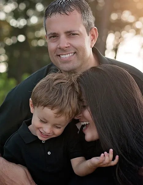 Dr. Daniel Lawless outdoors with his family, smiling, his young son in his arms.